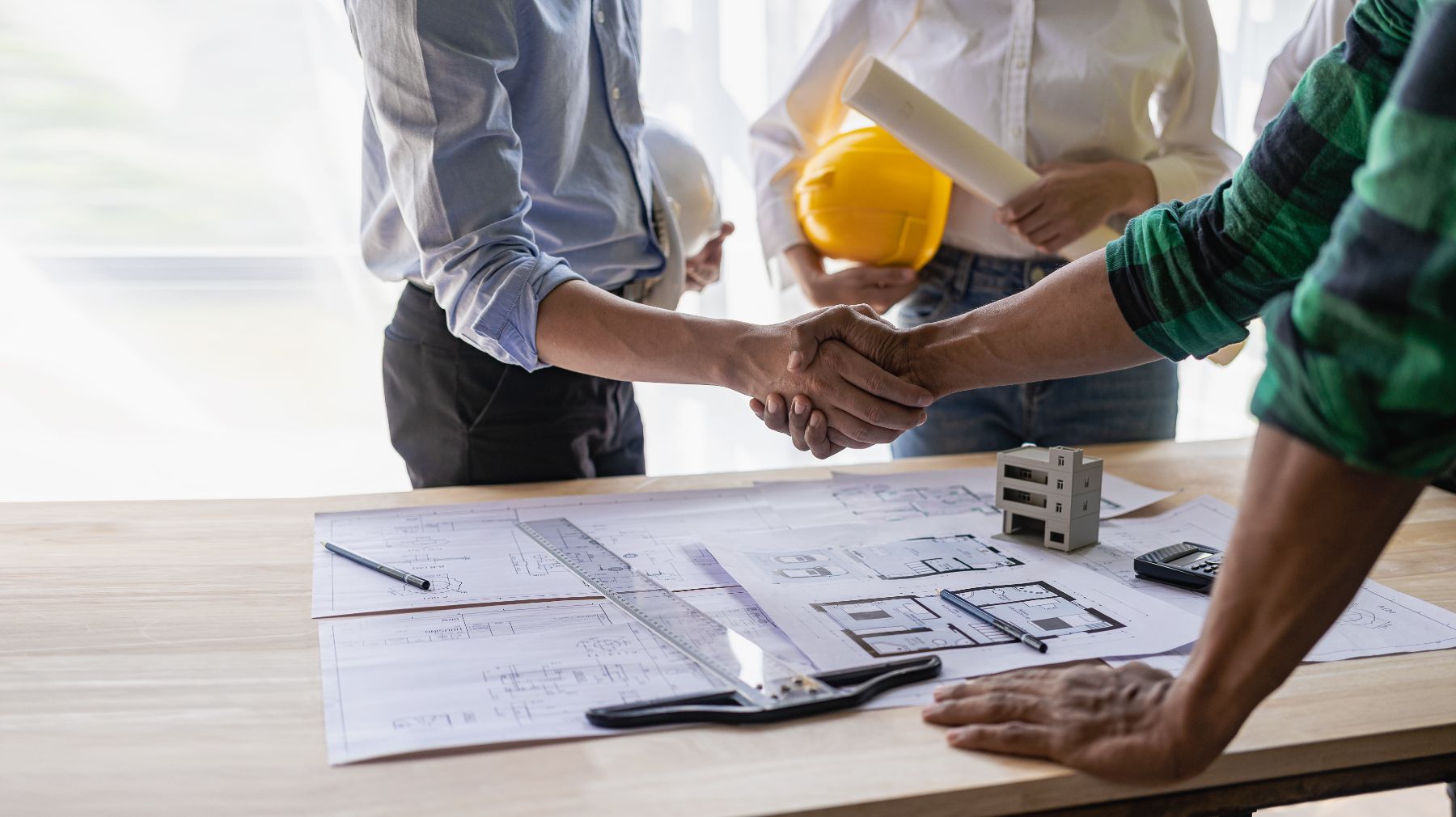 Construction workers shaking hands over blueprints and yellow hard hat at table