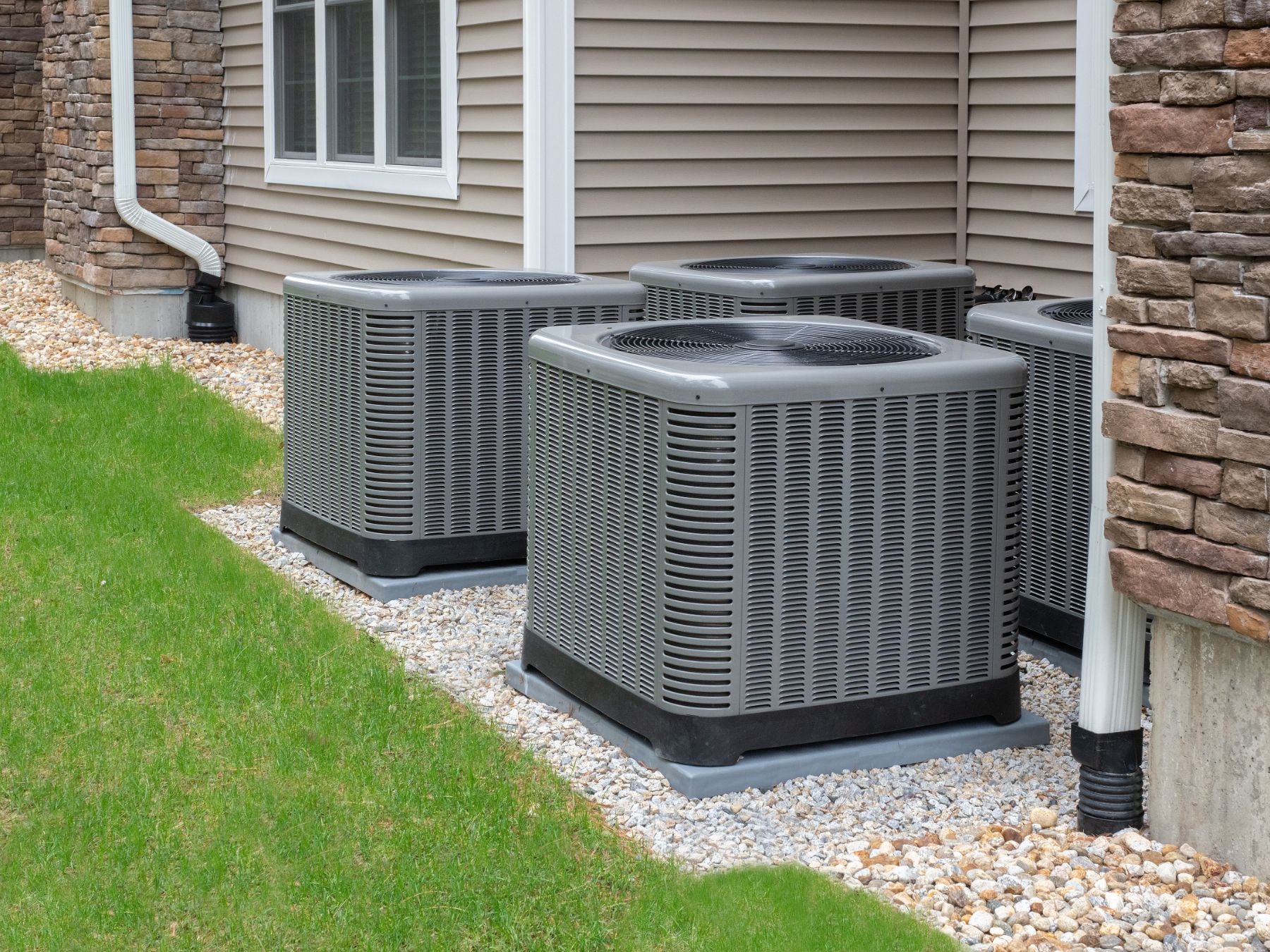 Multiple gray HVAC air conditioning units installed on gravel beside residential home