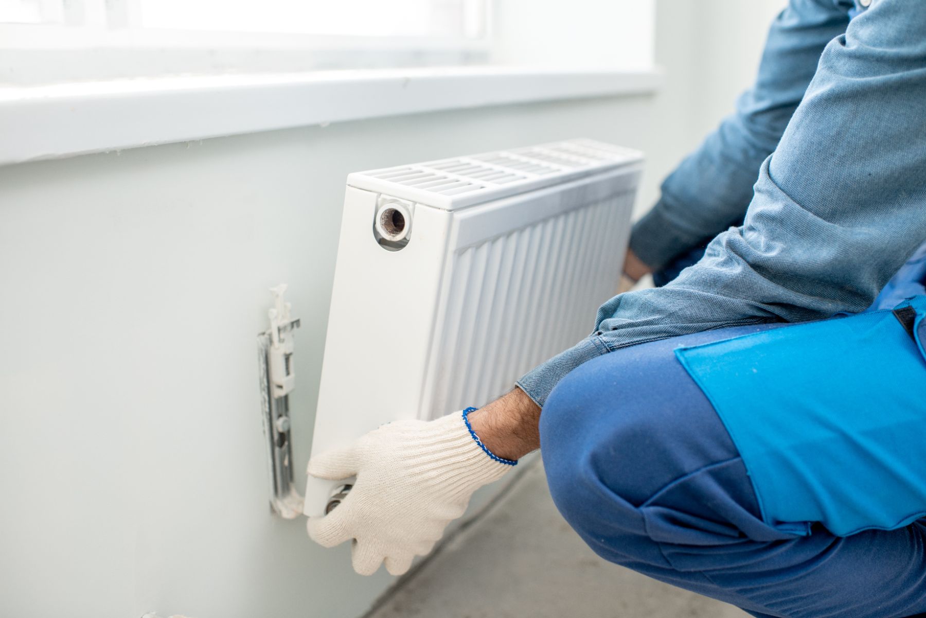 Technician in blue jacket and white gloves installing wall-mounted radiator near window