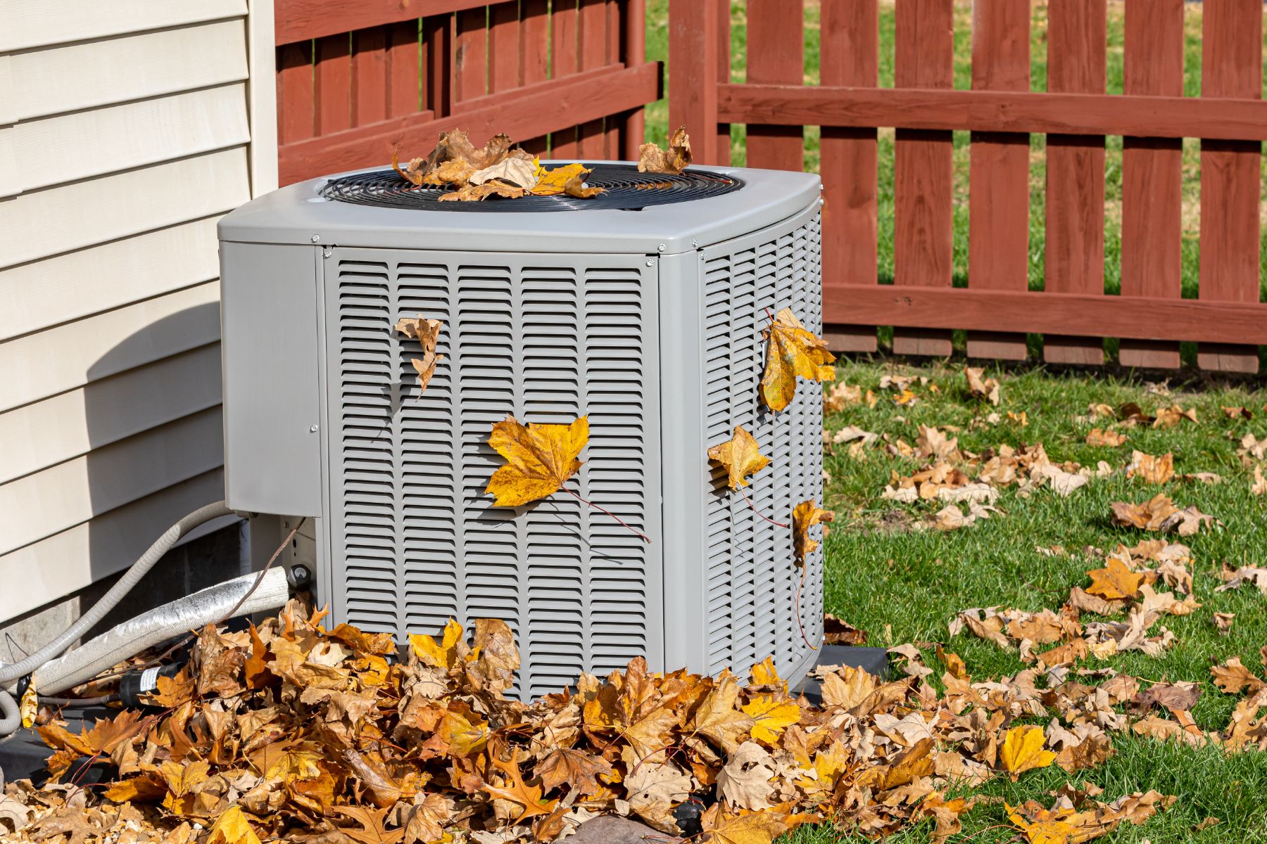 Gray HVAC outdoor unit surrounded by autumn leaves in a residential backyard