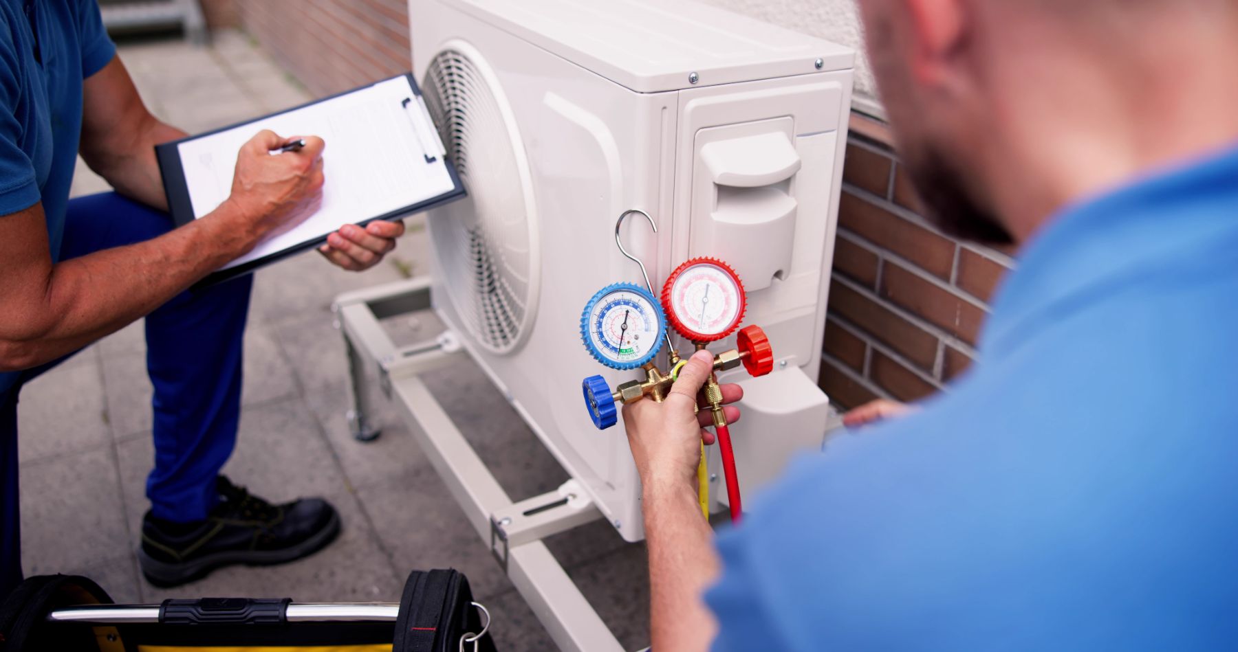 HVAC technician using manifold gauges to service air conditioning unit while colleague records data on clipboard