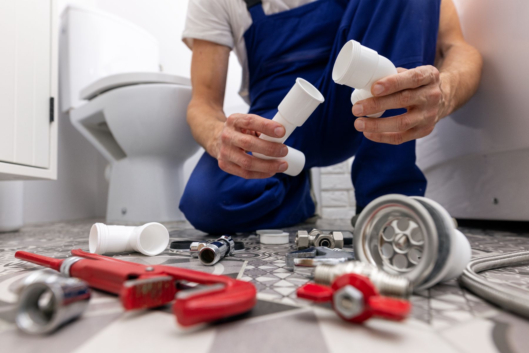 Plumber in blue uniform holding PVC pipes with tools and fittings on floor near toilet
