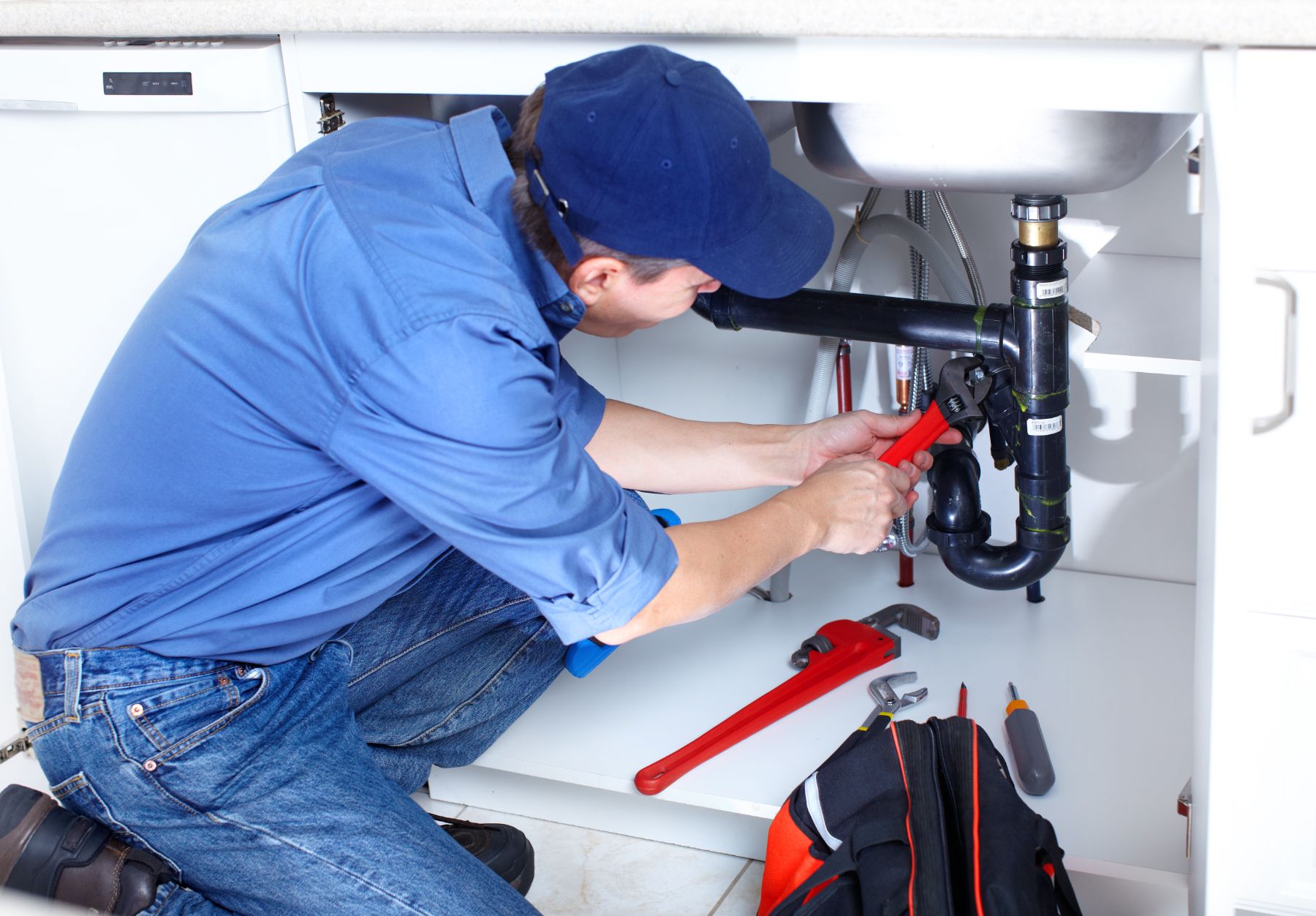 Plumber in blue uniform and cap working on under-sink pipes with red wrench