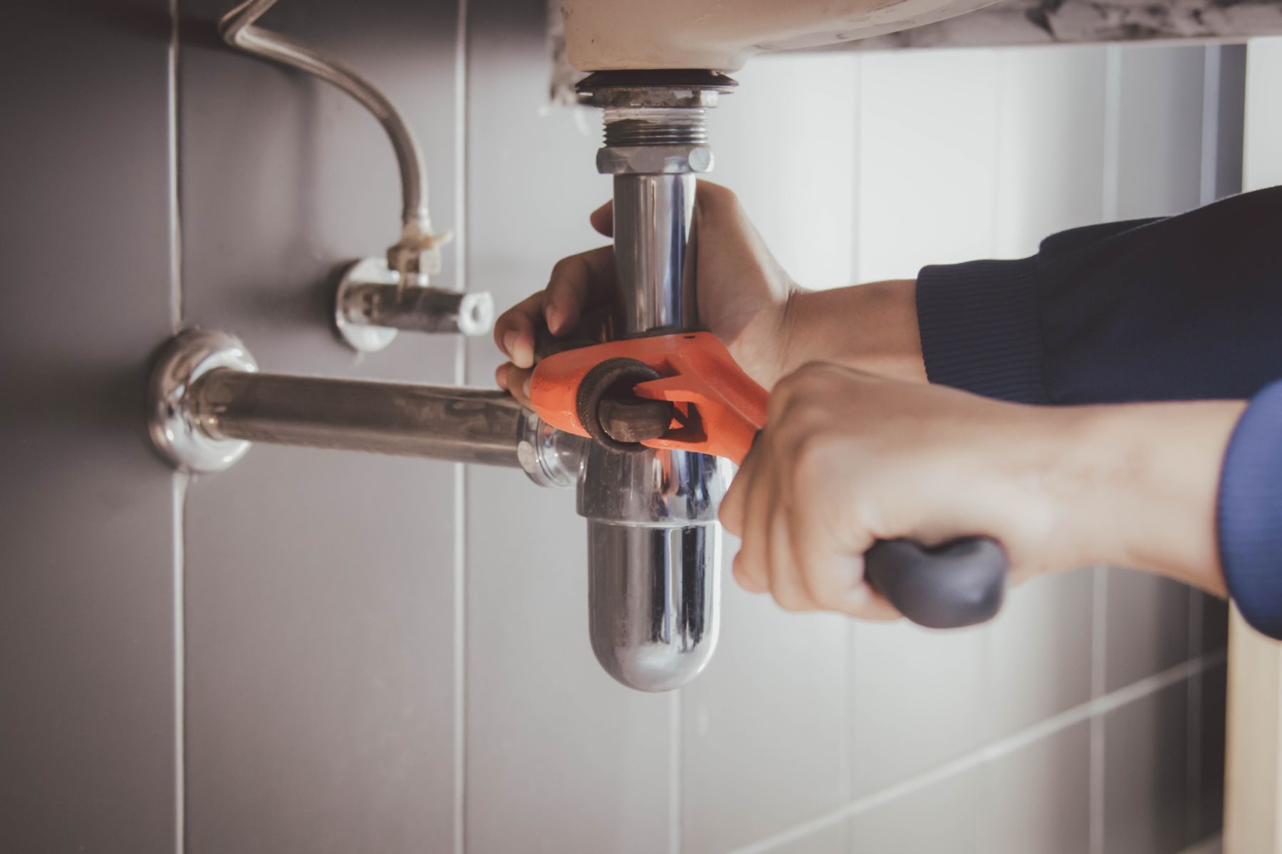 Plumber in orange gloves installing chrome P-trap under sink with wrench